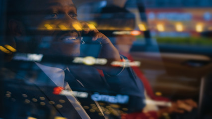 Wall Mural - Handsome Businessman and Beautiful Businesswoman Riding on the Backseat of a Car in the Evening. Man Makes a Phone Call, Woman Works on a Laptop. Camera Shot made from Outside the Car.