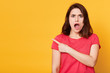 Portrait of astonished woman stands isolated on yellow studio background. Female wears red t shirt, looks at camera with open mouth, points her fore finger aside. Facial expression concept. Copy space