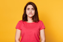 Portrait Of Young Pretty Emotional Woman On Yellow Background, Bites Her Lip, Looks At Camera With Scared Facial Expression, Afraids Of Something, Warches Horror Film. People And Emotions Concept.