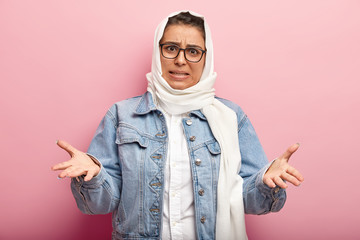 Wall Mural - Studio shot of annoyed emotional woman wears modern Muslim clothes and white headdress, spreads hands with displeasure, has specific appearance, follows Islamic religion. People and faith concept