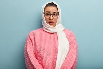 Wall Mural - Headshot of puzzled confused Muslim woman looks with indignation, hears shocking news, focused directly at camera, wears winter sweater, covers head with hijab, isolated over blue background