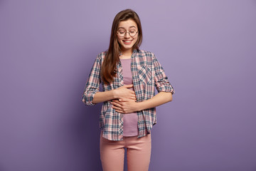Wall Mural - Glad woman has full stomach after delicious dinner, keeps both hands on belly, being well fed smiles positively dressed in casual clothes round spectacles isolated over purple wall. Feeling of satiety