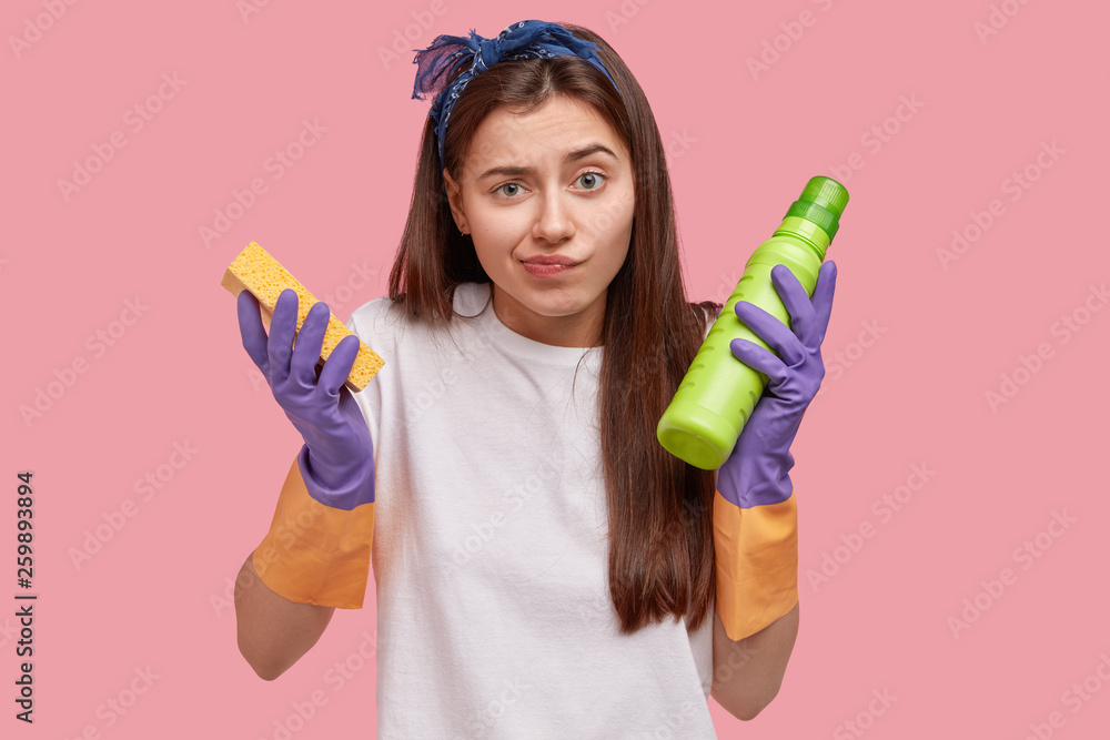 Housekeeping and cleaning concept. Unhappy confused young female janitor with long hair, holds sponge and green bottle with detergent, wears rubber gloves for hand protection, does house chores