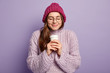 © wayhome.studio  - Beautiful satisfied European woman keeps eyes closed, holds coffee to go, warms with hot drink, wears pink headgear, knitted jumper, isolated over purple background. Drinking and spare time concept