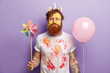 Studio shot of dissatisfied redhead man holds toy windmill and helium balloon, has face dirty with yellow watercolours, ginger hair and beard, isolated over purple background. Party preparation