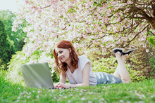 Young Smilling Hipster Redhead Girl Working On Her Laptop In Blossom Cherry Trees Garden. Blogger Preparing Posts For Social Media In The Garden Of Cherry Blossoms. Spring Time. Template. Blank. 