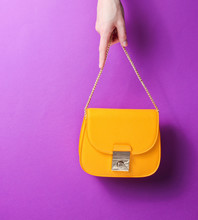 Female Hand Hold Fashionable Yellow Leather Bag With Golden Chain On Purple Background.