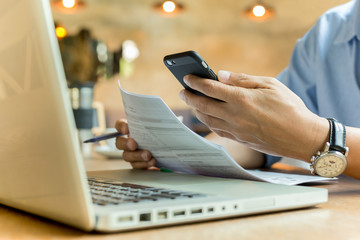 Business man using cell phone and holding document with laptop on table.