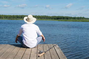 man in a hat sits on a pier with a fishing rod and looks at the river