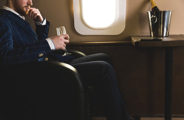 A young successful businessman in an expensive suit sits in the chair of a private jet with a glass of champagne in his hand and looks out the window