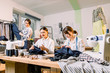 © sofiko14 - Female fashion designer standing near clothing rack with fashionable stylish handmade clothes. Two young girls dressmakers working on new pants collection for clients in cozy workshop studio