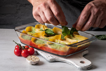 Cooking by chef hands traditional pasta recipe with greens, herbs, tomatoes and cheese, baked in glass tray on white wooden background.