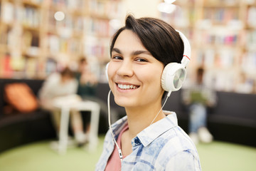 Wall Mural - Smiling beautiful hipster girl listening to audiobook in headphones and looking at camera while spending time in library