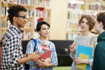 Wall Mural - Group of content pensive young multiethnic university students standing in modern library area and talking while planning project together