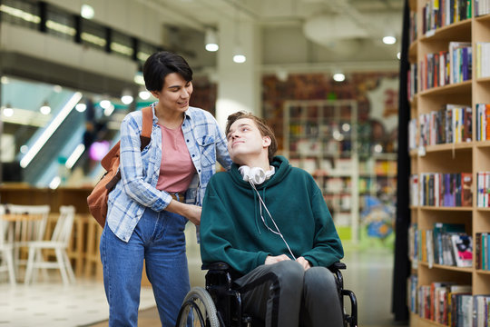 positive attractive young woman with satchel pushing wheelchair and talking to disabled student in l