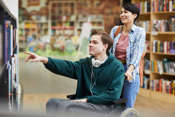 Content handsome boy with wired headphones on neck sitting in wheelchair and pointing at bookshelf while choosing book with nurse