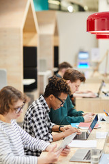 Wall Mural - Group of concentrated multiethnic students sitting in line and doing hometask in library, focus on African guy typing on laptop