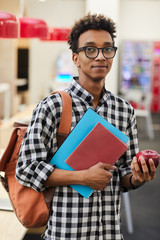 Wall Mural - Content young African-American student in glasses wearing checkered shirt and satchel standing in library area and holding ripe apple