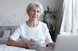 Attractive confident female pensioner wearing white blouse and pearl necklace drinking coffee or tea, looking through papers at cozy home office, using wireless internet conneciton on laptop