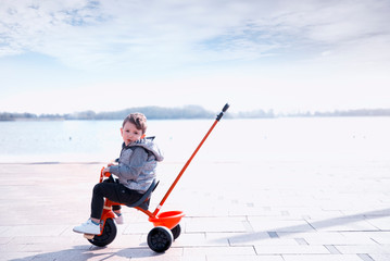 A little happy boy rides on red tricycle on a  street looking at camera