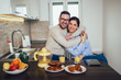 © Mediteraneo - Happy young couple in their kitchen posing with their heads close together behind the counter smiling at the camera