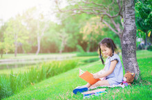Cute Little Girl Reading A Book With A Doll In The Park