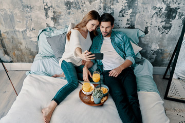 Sticker - Let me take a selfie! Top view of beautiful young couple taking selfie and smiling while spending time in bed at home