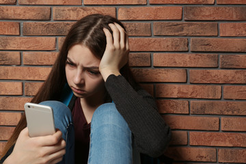 Wall Mural - Upset teenage girl with smartphone sitting at wall indoors. Space for text
