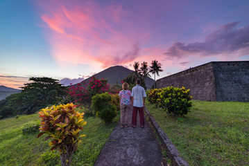 Wall Mural - Api volcano at sunset, couple looking at view from Banda Naira fort, Maluku Moluccas Indonesia, Top travel tourist destination, dramatic sky.