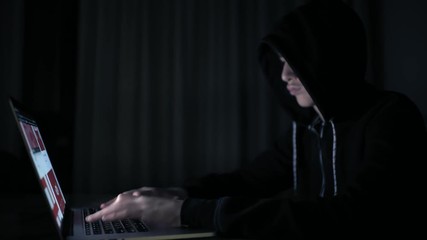 Hacker's hacking into the computer network. A young guy in a dark room typing on a laptop.  Hacker with a laptop. E-the robber. Lonely teen chatting online.