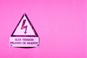 Pink 'high voltage and risk of death' sign in Spanish on a plastic pink painted plastered wall.