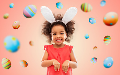 Wall Mural - childhood, party props and easter concept - happy little african american girl wearing bunny ears headband over colored eggs on living coral background