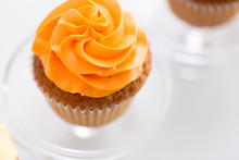 Food, Pastry And Sweets Concept - Cupcake With Orange Buttercream Frosting On Glass Confectionery Stand Over White Background