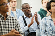 © Seventyfour - Content handsome mature African conference participant in glasses wearing suspenders sitting in audience and applauding for speaker while looking at camera