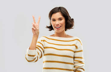 gesture, counting and people concept - happy smiling young woman in striped pullover showing peace hand sign or two fingers over grey background