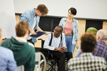 Live interaction with disabled young scientist: content young man in glasses supporting black man in wheelchair while talking to him in front of audience at conference