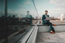 Young Smiling Cheerful Caucasian Bearded Businessman In Formal Wear Using Laptop While Sitting On Rooftop. At First They Will Ask You Why You Are Doing It, Later They Will Ask You How You Did It.