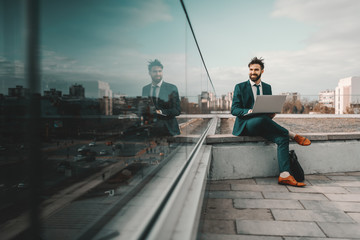 Young smiling cheerful Caucasian bearded businessman in formal wear using laptop while sitting on rooftop. At first they will ask you why you are doing it, later they will ask you how you did it.