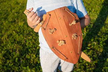 Young white kid playing with wooden toys outside in countryside meadow on sunny sunset summer evening. Boy holds sword and shield. Horizontal color photography.