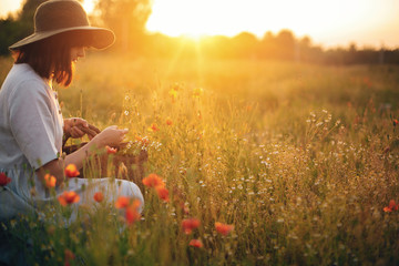 Wall Mural - Stylish girl in linen dress gathering flowers in rustic straw basket, sitting in poppy meadow in sunset. Boho woman in hat relaxing in warm evening sunlight in summer field. Space for text