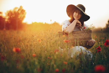 Wall Mural - Stylish girl in linen dress smiling in poppy meadow in sunset light with flowers in rustic straw basket. Boho woman in hat relaxing in summer field. Atmospheric moment, space text