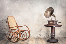 Vintage Antique Gramophone Phonograph Turntable With Brass Horn On Wooden Table And Aged Rocking Chair Front Concrete Wall Background With Shadow. Retro Old Style Filtered Photo 