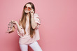 Beautiful and happy blonde model girl with a shiny smile in beige blouse and in fashionable pink sunglasses holding stylish shoes in her hands and posing at the pink background, isolated