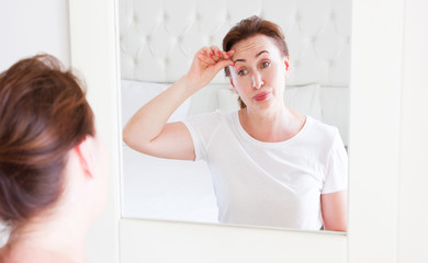 Wall Mural - Middle age woman looking in mirror on face wrinkle forehead in bedroom. Wrinkles and anti aging skin care concept. Selective focus. Banner