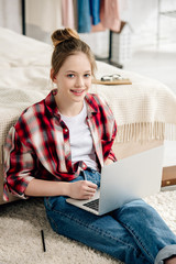 Wall Mural - Smiling teenager in jeans sitting on carpet and using laptop