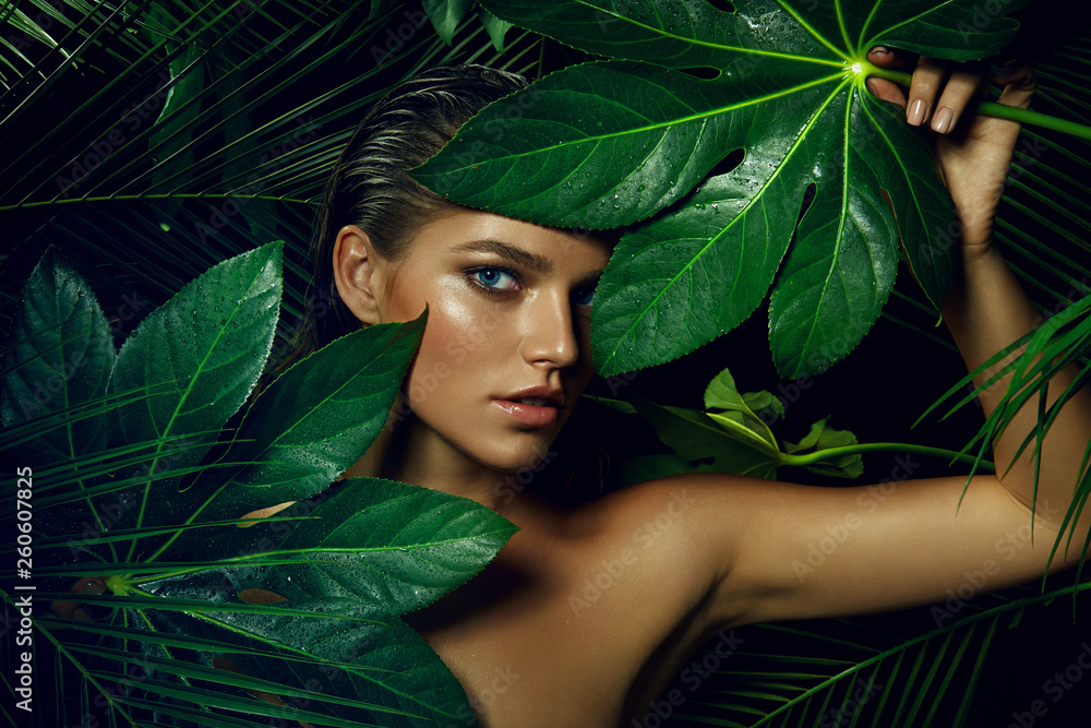 A beautiful tanned girl with natural make-up and wet hair stands in the jungle among exotic plants. - obrazy, fototapety, plakaty 
