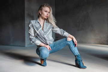 Wall Mural - Fashionable modern young woman blonde in stylish shirt in trendy torn jeans in vintage cowboy boots posing sitting in a room with sunlight. American beautiful fashion model girl. Modern women's style.