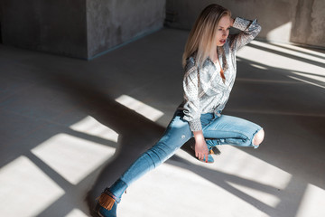 Wall Mural - American young blonde woman in stylish shirt in vintage ripped jeans in trendy cowboy boots posing sitting indoors with sunlight. Sexy beautiful fashion model girl relaxes in a sunny room.