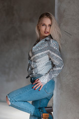 Wall Mural - Modern beautiful young woman blonde with sexy lips with blond hair in ripped stylish jeans in a fashionable shirt in green shoes posing near a gray wall. Attractive girl relaxes indoors. Fashion.