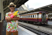 Happy Elderly Traveler Asian Man Wearing Summer Shirt, Straw Hat And Sunglasses Holding Colorful Water Squirt Gun Over Old Diesel Train And Platform Of Railway Station, Summer Water Festival Holiday C